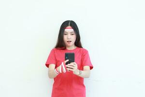 portrait of beautiful asian woman wearing red outfit celebrating Indonesia independence day holding mobile phone with surprised expression photo