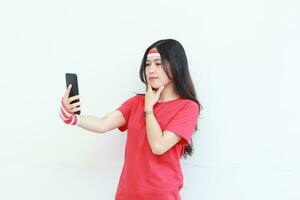 portrait of beautiful asian woman wearing red outfit celebrating Indonesia independence day holding mobile phone with thinking expression photo