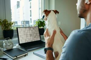 Man sitting at table by the window with dog and using laptop photo