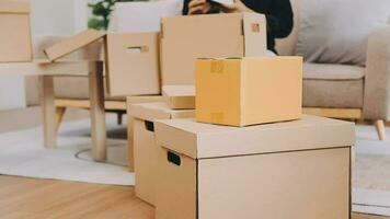 Two people couple interracial family ameste in living room in new apartment open cardboard boxes receive parcels with orders from internet store with new interior items rejoice at courier delivery video