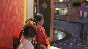 Cheerful Asian family at cinema show. Mum and kids sit in theater seat, waiting for father's popcorn before watching movie, and happy together, a public indoor entertainment lifestyle with film shows. video