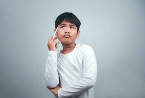 un chico en un blanco camisa es participación un blanco bolígrafo en un blanco antecedentes. muestra pensamiento, reflexionando, y considerando opciones foto