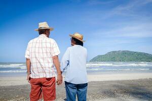 An elderly Asian couple Stand hand in hand on the beach Look at the beautiful sea in the morning together. Travel concept to live happily in retirement age. copy space photo