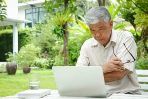 Asian elderly man Use a laptop to sit and work in the garden. in the residential area He holds eyeglasses and uses his thoughts to work. concept of health care for seniors in retirement age photo