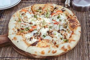 Various ingredients are served in a pizza such as mozzarella cheese, onions, red peppers, green peppers, mushrooms, shredded chicken, oregano leaves. Served on a tray made of whole wood. photo