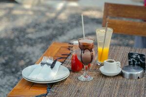 A dining table with cold chocolate and orange squash drinks served in a glass with a stainless steel straw. Accompanied by 2 bottles of ketchup, a serving plate and a glass of liquid sugar. photo