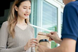 Happy smiling Asian woman receives paper box parcel of food from courier front house. Delivery man send deliver express. online shopping, paper container, takeaway, postman, delivery service, packages photo