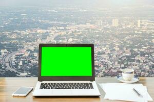 Bosquejo imagen de ordenador portátil con blanco verde pantalla, teléfono inteligente, café taza en de madera mesa ver al aire libre de oficina edificio denso paisaje urbano alto ángulo ver antecedentes. foto