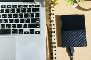 externo difícil disco en cuaderno con ordenador portátil teclado con un lápiz y flor maceta árbol en de madera fondo, arriba ver oficina mesa. foto