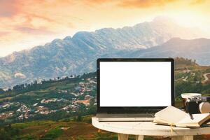 Bosquejo imagen de ordenador portátil con vacío pantalla con cámara, cuaderno, café taza en mesa con a montañas paisaje en contra puesta de sol cielo fondo,ocio y viaje concepto. foto