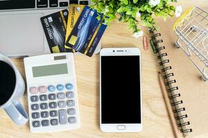 crédito tarjeta en compras carro con portátil, cuaderno, flor maceta árbol, teléfono inteligente y calculadora en de madera fondo, en línea bancario concepto,arriba ver oficina mesa. foto