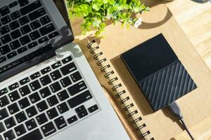 externo difícil disco en cuaderno con ordenador portátil teclado con un lápiz y flor maceta árbol en de madera fondo, arriba ver oficina mesa. foto