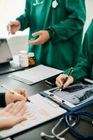 Medical team having a meeting with doctors in white lab coats and surgical scrubs seated at a table discussing a patients working online using computers in the medical photo