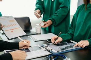 Medical team having a meeting with doctors in white lab coats and surgical scrubs seated at a table discussing a patients working online using computers in the medical photo