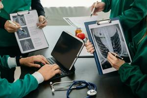 Medical team having a meeting with doctors in white lab coats and surgical scrubs seated at a table discussing a patients working online using computers in the medical photo