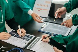 Medical team having a meeting with doctors in white lab coats and surgical scrubs seated at a table discussing a patients working online using computers in the medical photo