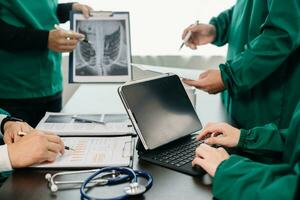 Medical team having a meeting with doctors in white lab coats and surgical scrubs seated at a table discussing a patients working online using computers in the medical photo