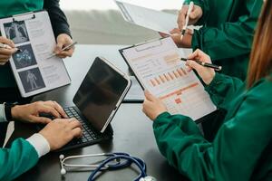 Medical team having a meeting with doctors in white lab coats and surgical scrubs seated at a table discussing a patients working online using computers in the medical photo