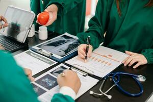 Medical team having a meeting with doctors in white lab coats and surgical scrubs seated at a table discussing a patients working online using computers in the medical photo