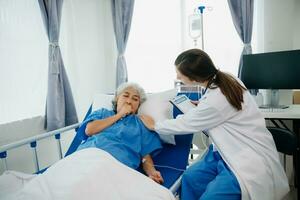 médico en blanco traje tomar notas mientras que se discute y asiático anciano, mujer paciente quien acostado en cama con recepción salina solución en hospital o clínica. foto