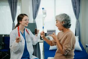 Doctor in white suit take notes while discussing and Asian elderly, woman patient who lying on bed with receiving saline solution in hospital or clinic. photo