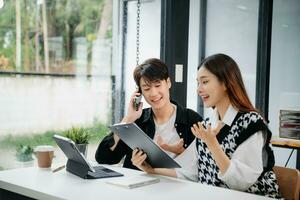 Asian business colleague business presents and explains and using laptop and tablet. Teamwork, financial marketing team,   while sitting in modern office room. photo