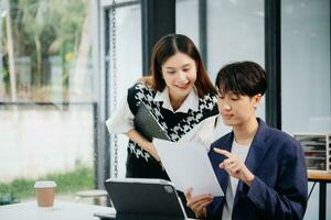 Asian business colleague business presents and explains and using laptop and tablet. Teamwork, financial marketing team,   while sitting in modern office room. photo