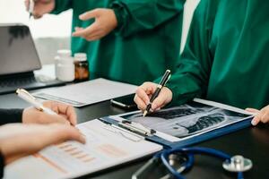 Medical team having a meeting with doctors in white lab coats and surgical scrubs seated at a table discussing a patients working online using computers in the medical photo