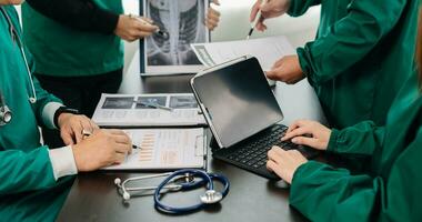 Medical team having a meeting with doctors in white lab coats and surgical scrubs seated at a table discussing a patients working online using computers in the medical photo