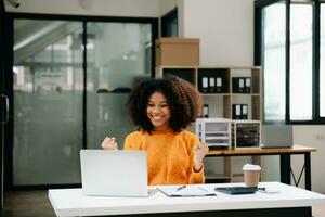 Young attractive Asian woman smiling thinking planning writing in notebook, tablet and laptop working from home, looking at camera at office photo