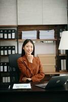 Young attractive Asian woman smiling thinking planning writing in notebook, tablet and laptop working from home, looking at camera at office photo