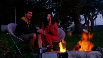 couple dans l'amour est séance par le Extérieur feu de camp dans le Cour de le maison sur camping chaises, une romantique soir. une homme et une femme chaud leur mains par le feu, boisson thé video