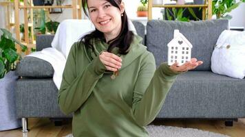 A happy woman in her house holds in her hands a miniature figure of a house in the interior. Dream house project, real estate purchase, insurance, mortgage, rent, reservation video