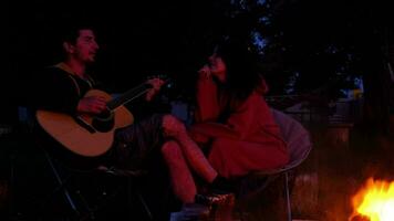 A man plays the guitar, a woman listens and sings along. A couple in love is sitting by the outdoor campfire in the courtyard of the house on camping chairs, a romantic evening video