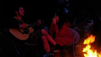 une homme pièces le guitare, une femme écoute et chante le long de. une couple dans l'amour est séance par le Extérieur feu de camp dans le Cour de le maison sur camping chaises, une romantique soir video