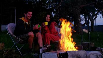 couple dans l'amour est séance par le Extérieur feu de camp dans le Cour de le maison sur camping chaises, une romantique soir. une homme et une femme chaud leur mains par le feu, boisson thé video