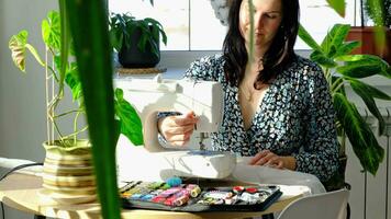 A woman sews tulle on an electric sewing machine in a white modern interior of a house with large windows, house plants. Comfort in the house, a housewife's hobby video