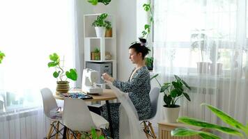 un mujer cose tul en un eléctrico de coser máquina en un blanco moderno interior de un casa con grande ventanas, casa plantas. comodidad en el casa, un ama de casa pasatiempo video