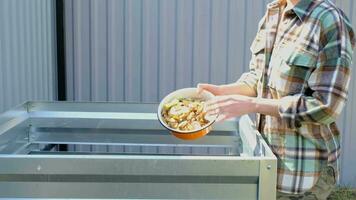 A woman in a plaid shirt pours food waste from a bowl into a compost heap of potato and carrot peelings. Compost box made of metal, eco-friendly fertilizer for the garden video