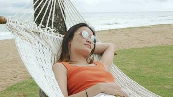 asiático mujer relajante y en hamaca cerca playa debajo Coco árbol video
