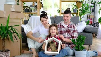 Moved family with a child and a cat pet are enjoying a new home, sitting on the sofa and holding the roof near boxes with things. Mortgage, insurance, buying and moving into your housing video