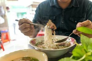 A man using chopsticks and spoon eating traditional Pho Bo vietnamese soup with beef and rice noodles on a metal table, real scene in local restaurant photo