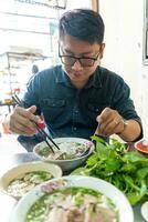 un hombre utilizando palillos y cuchara comiendo tradicional pho bo vietnamita sopa con carne de vaca y arroz tallarines en un metal mesa, real escena en local restaurante foto