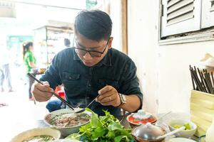 un hombre utilizando palillos y cuchara comiendo tradicional pho bo vietnamita sopa con carne de vaca y arroz tallarines en un metal mesa, real escena en local restaurante foto