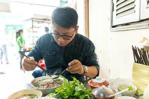 A man using chopsticks and spoon eating traditional Pho Bo vietnamese soup with beef and rice noodles on a metal table, real scene in local restaurant photo