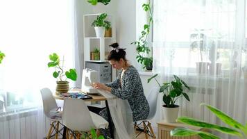 A woman sews tulle on an electric sewing machine in a white modern interior of a house with large windows, house plants. Comfort in the house, a housewife's hobby video