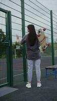 meisje in overmaats kleren op zoek Bij een basketbal rechtbank met een longboard in haar handen, verticaal video