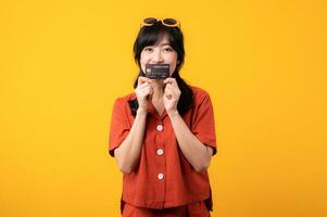 Portrait young beautiful asian woman happy smile dressed in orange clothes showing credit card isolated on yellow background. Pay and purchase shopping payment concept. photo