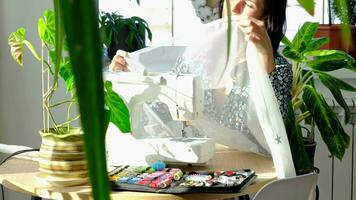 un mujer cose tul en un eléctrico de coser máquina en un blanco moderno interior de un casa con grande ventanas, casa plantas. comodidad en el casa, un ama de casa pasatiempo video