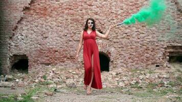 magnifique sexy femme dans une rouge soir robe avec impressionnant maquillage sur Halloween va avec coloré fumée contre le toile de fond un abandonné bâtiment. peinture à la graisse pour le vacances sur octobre 31. lent mouvement video